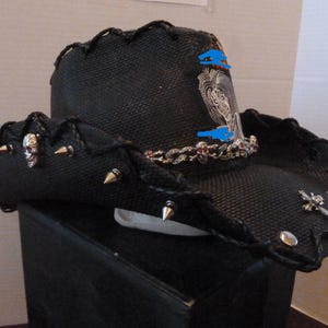 May include: Black cowboy hat with a woven texture, adorned with silver spikes, a skull, and a decorative chain. The hat features a scalloped brim and a silver sword charm, creating a bold, edgy aesthetic.