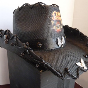 May include: Black cowboy hat with a woven texture, adorned with a leather band and decorative metal accents. Features the "Ozzy Osbourne" logo and skull and bat embellishments. The brim is trimmed with braided detailing and metal studs.