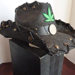 May include: A black woven cowboy hat featuring a green cannabis leaf patch on the front, a black leather band with a circular metal emblem, bronze-toned skull studs along the brim, and the word Peace in metal lettering on the front brim.