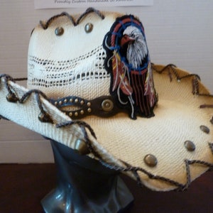 May include: Cream-coloured cowboy hat made of woven straw. It has a leather band with studs and a patch showing an eagle with a dreamcatcher. The brim is trimmed with dark brown stitching and metal studs. A Western-style hat.