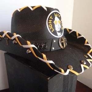 Op de afbeelding: Zwarte cowboyhoed met witte en gele stiksels rond de rand. De hoed heeft een leren band met een kruisvormige gesp en een ronde patch met de tekst "LIGHT MY FIRE" en een afbeelding van een man met een zonnebril.