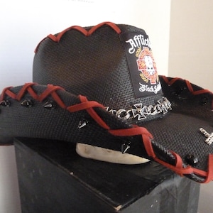May include: A black cowboy hat featuring a textured finish, accented with red lacing along the brim and crown. The hat includes black conical studs, a silver-toned chain band with cross emblems, and a patch displaying the text Affliction Black.
