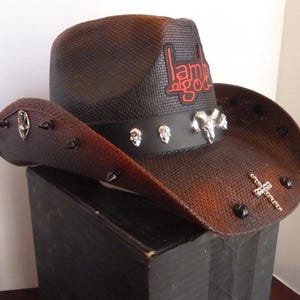 Peut inclure: Un chapeau de cowboy marron et noir avec le texte "Lamb of God" en rouge. Le chapeau présente un bandeau en cuir noir avec des crânes argentés et un crâne de bélier. Le bord est orné de pointes et d'une croix.