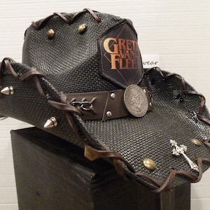 May include: A black and brown cowboy hat with a leather band and metal studs. The hat has a patch with the text "Gretchen Van Fleet" on it.