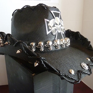Puede incluir: Sombrero de vaquero negro con detalles de calaveras plateadas, púas y un diseño de cruz. El sombrero presenta una textura tejida y una banda decorativa de pequeñas calaveras alrededor del borde. Un accesorio único.