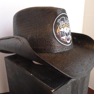 Peut inclure: Chapeau de cowboy en paille noire avec un large bord. Le chapeau présente un écusson circulaire avec le texte "Von Dutch" au-dessus d'un motif de crâne et de flammes. Le chapeau est présenté sur une surface noire.