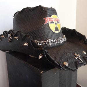 May include: Black cowboy hat with silver studs and a decorative chain. The hat features the word "Megadeath" in red and yellow. A unique accessory.