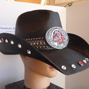 May include: Black cowboy hat with a decorative band and a large silver and rose-colored emblem. The hat brim is adorned with clear and red rhinestones. The hat is displayed on a mannequin head.