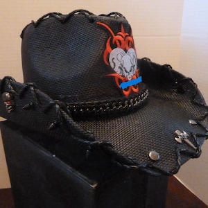 Peut inclure: Chapeau de cowboy noir avec un bord décoratif, orné d'un motif de crâne et de bélier avec des accents rouges et orange. Le chapeau a des détails de chaîne et de pointes, et une ligne bleue.