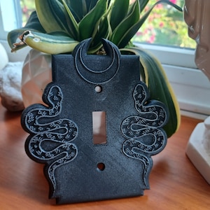 May include: Black light switch cover with a crescent moon and two snakes on either side. The snakes are detailed with scales and are in a curled position.