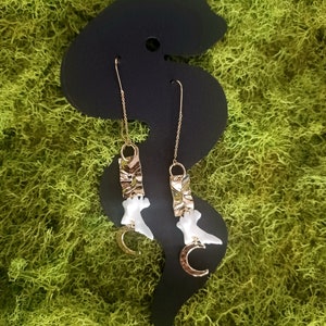 May include: A pair of gold earrings with white ceramic crescent moon charms and textured gold accents hanging from a black display card.