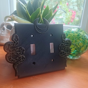 May include: Black light switch cover with a crescent moon and snake design. The switch cover is made of 3D printed plastic.