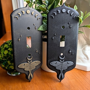 May include: Two black light switch covers with celestial designs. Each cover features a moth, crescent moons, and stars. One cover has gold accents, the other silver. The covers are decorative and add a unique touch to any room.