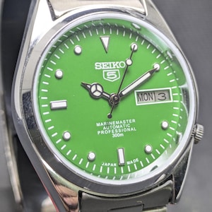 Könnte beinhalten: Eine Seiko 5 Marine Master Automatik-Professional-Uhr aus Edelstahl mit einem leuchtend grünen Zifferblatt. Die Uhr verfügt über eine Tages- und Datumsanzeige, Leuchtzeiger und den Text "Marine Master Automatic Professional 300m".