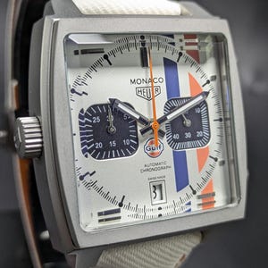Op de afbeelding: Een vierkant Heuer Monaco chronograaf horloge met een witte wijzerplaat, blauwe en oranje racestrepen, twee donkere sub-wijzerplaten, een feloranje centrale wijzer en een datumvenster op de zes-uur positie. De wijzerplaat bevat het Gulf logo.