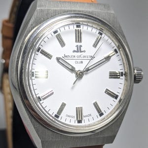 Op de afbeelding: Een zilverkleurig Jaeger-LeCoultre Club horloge met een witte wijzerplaat en een bruine leren band. Het horloge heeft een rechthoekige kast, zilveren wijzers en de tekst "JAEGER-LECOULTRE CLUB" op de wijzerplaat. Het horloge is gemarkeerd met "SWISS MADE".