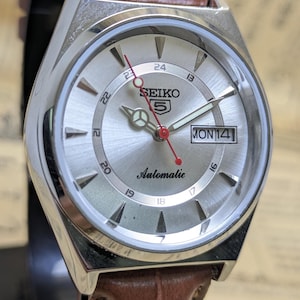 Puede incluir: Un reloj de pulsera automático Seiko 5 plateado con una correa de cuero marrón. La esfera del reloj es plateada con números y agujas negras, y un segundero rojo. El día y la fecha se muestran a las 3 en punto.