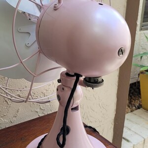 Puede incluir: Un ventilador el&eacute;ctrico rosa de estilo vintage con aspas blancas. El ventilador tiene una carcasa de motor redondeada, un asa y una base decorativa. El ventilador est&aacute; sobre una mesa de madera. La jaula del ventilador es de color rosa claro.