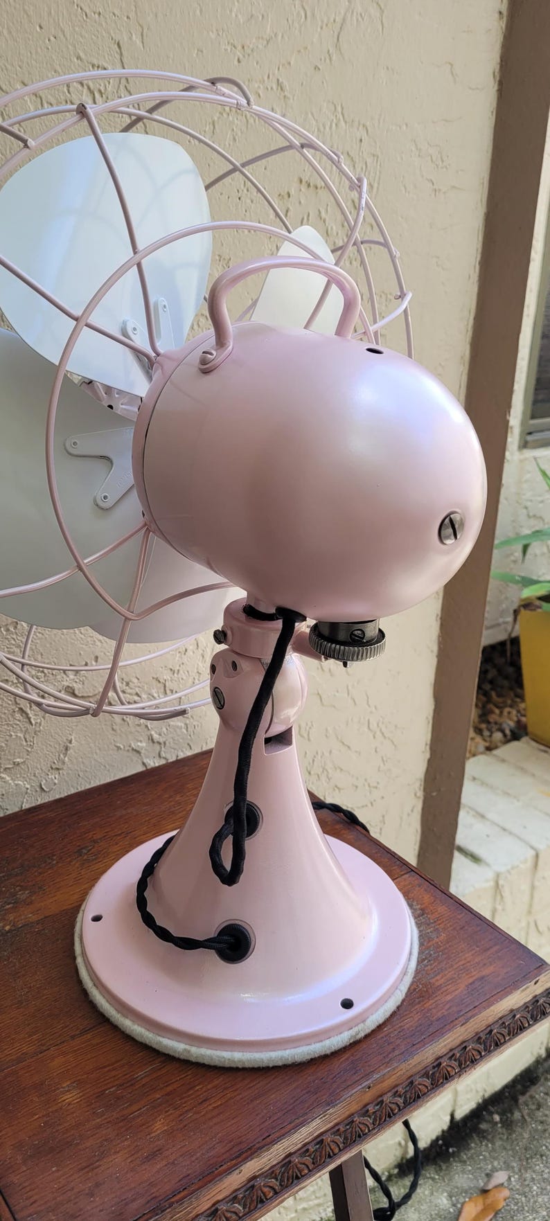 Puede incluir: Un ventilador el&eacute;ctrico rosa de estilo vintage con aspas blancas. El ventilador tiene una carcasa de motor redondeada, un asa y una base decorativa. El ventilador est&aacute; sobre una mesa de madera. La jaula del ventilador es de color rosa claro.