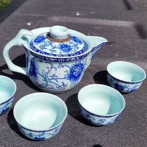 May include: A ceramic tea set with a teapot and four matching cups. The teapot and cups are light blue with a white floral pattern and blue accents. The teapot has a lid and a curved handle.