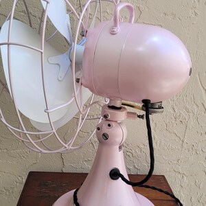 Puede incluir: Un ventilador el&eacute;ctrico rosa vintage con una aspa blanca y una jaula protectora. El ventilador est&aacute; sobre una mesa de madera. La base y la carcasa del motor del ventilador son de color rosa a juego, con un cable de alimentaci&oacute;n negro.