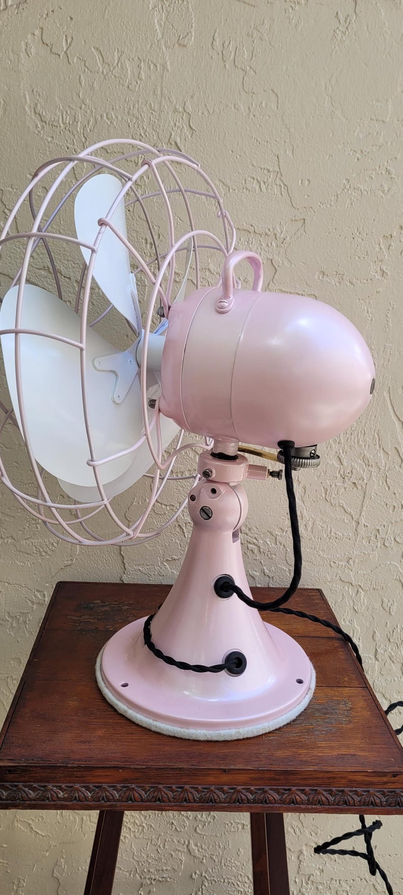 Puede incluir: Un ventilador el&eacute;ctrico rosa vintage con una aspa blanca y una jaula protectora. El ventilador est&aacute; sobre una mesa de madera. La base y la carcasa del motor del ventilador son de color rosa a juego, con un cable de alimentaci&oacute;n negro.