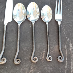 May include: A set of handcrafted silverware, including a knife, three spoons, and a fork. The utensils are made of silver-colored metal with a unique, curled design at the base of the handles. The silverware is arranged on a dark surface.