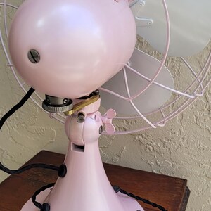 Puede incluir: Un ventilador el&eacute;ctrico rosa vintage con una jaula y aspas blancas. El ventilador tiene una carcasa de motor redondeada y una base de pedestal. El ventilador est&aacute; sobre una mesa de madera.