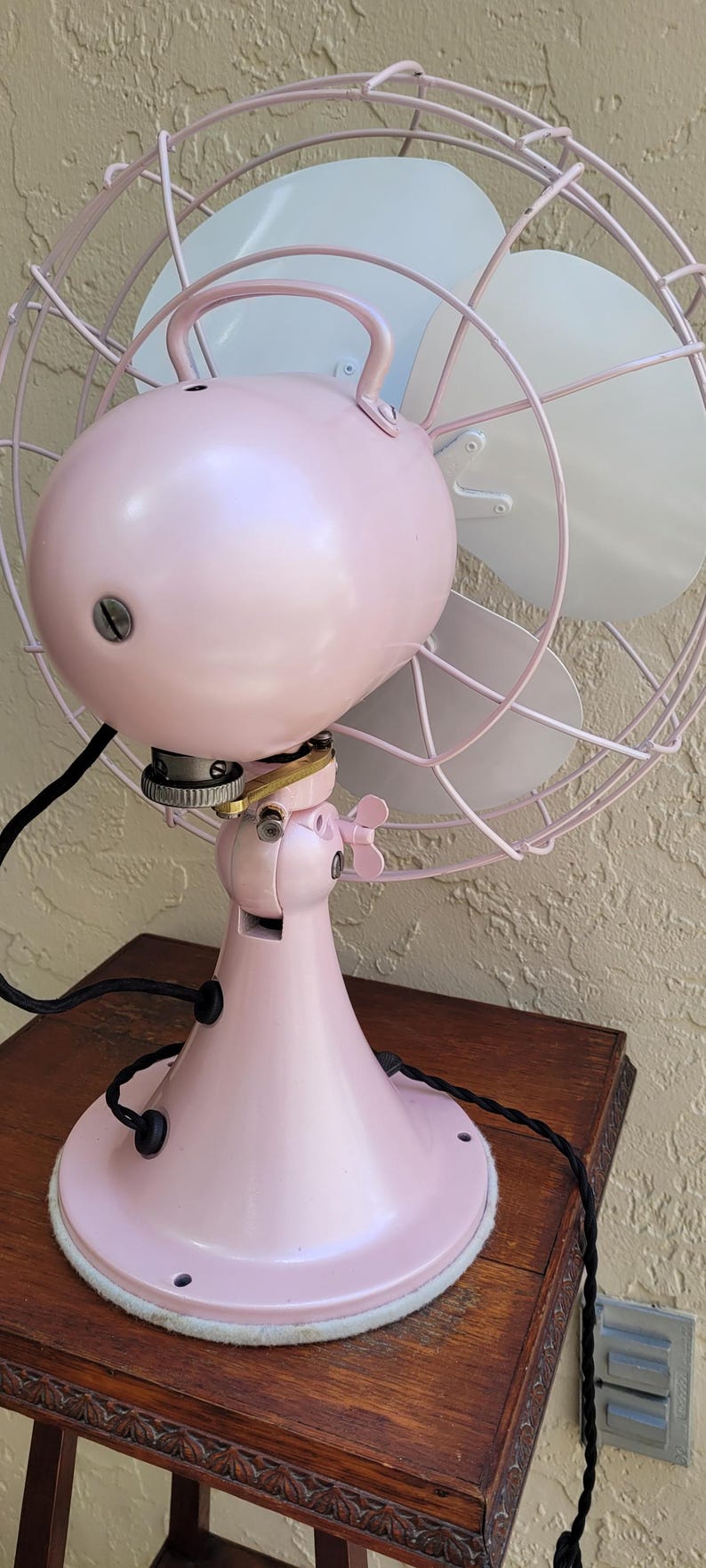 Puede incluir: Un ventilador el&eacute;ctrico rosa vintage con una jaula y aspas blancas. El ventilador tiene una carcasa de motor redondeada y una base de pedestal. El ventilador est&aacute; sobre una mesa de madera.