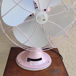 Puede incluir: Ventilador vintage Emerson Electric en un tono rosa suave, con cuatro aspas blancas y una jaula protectora. El ventilador est&aacute; sobre una base rosa a juego. El ventilador est&aacute; sobre una mesa de madera marr&oacute;n oscuro.