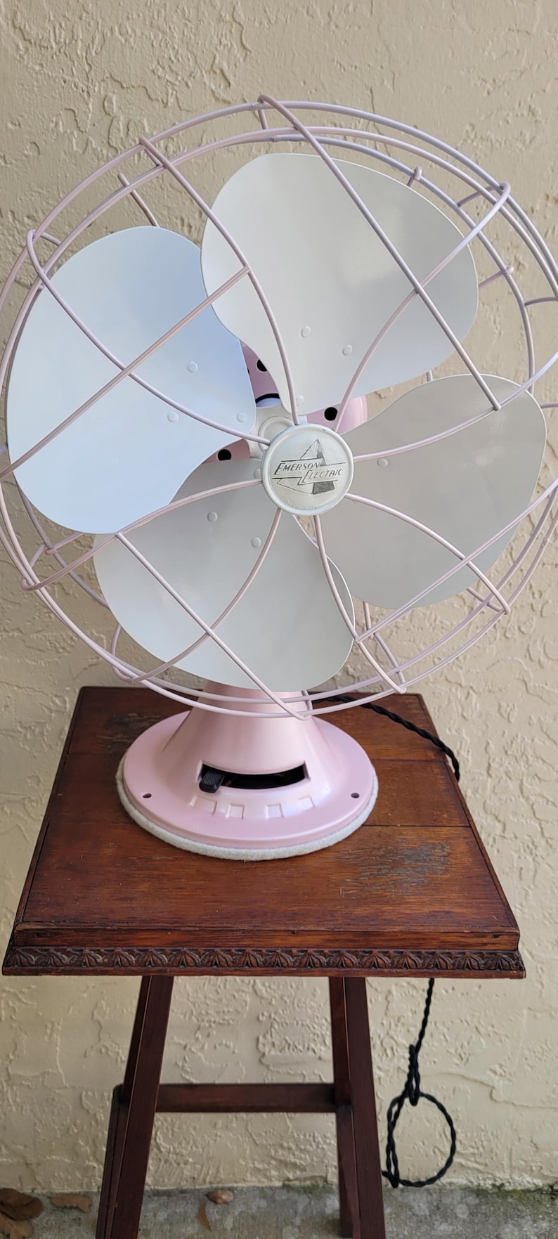 Puede incluir: Ventilador vintage Emerson Electric en un tono rosa suave, con cuatro aspas blancas y una jaula protectora. El ventilador est&aacute; sobre una base rosa a juego. El ventilador est&aacute; sobre una mesa de madera marr&oacute;n oscuro.