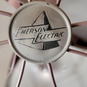 Puede incluir: Primer plano de un ventilador Emerson Electric. El ventilador tiene una jaula rosa claro y un emblema central con las palabras "Emerson Electric" y "St. Louis Mo".