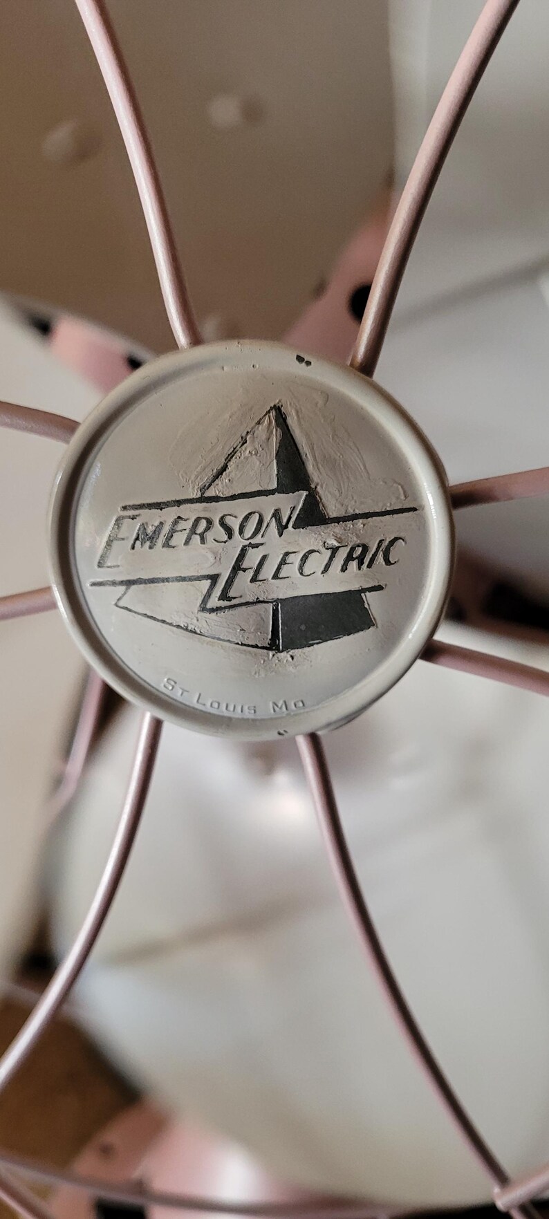 Puede incluir: Primer plano de un ventilador Emerson Electric. El ventilador tiene una jaula rosa claro y un emblema central con las palabras "Emerson Electric" y "St. Louis Mo".