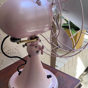 Puede incluir: Un ventilador el&eacute;ctrico rosa vintage con una jaula y aspas. El ventilador est&aacute; sobre una mesa de madera oscura. La carcasa del motor y la base del ventilador son de color rosa a juego. Las aspas son blancas.