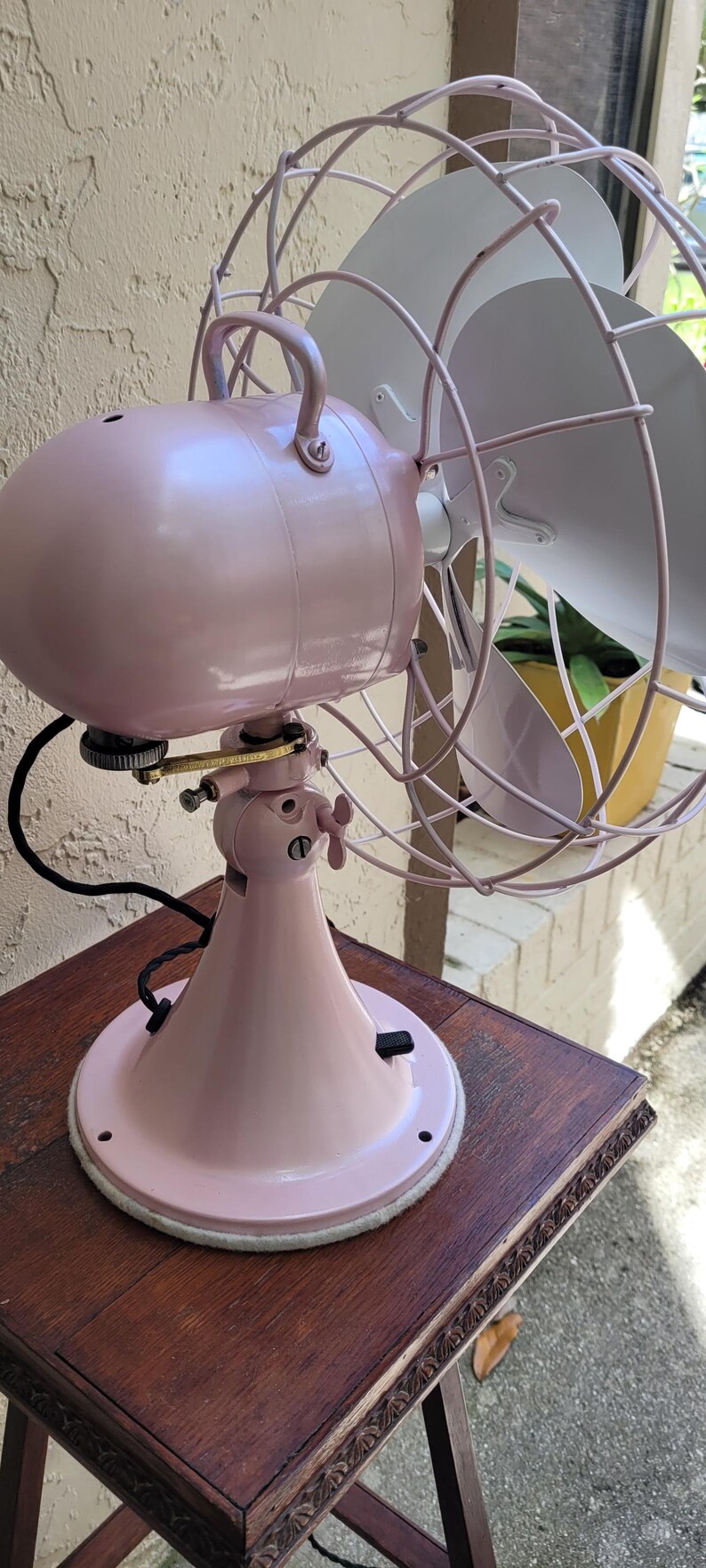 Puede incluir: Un ventilador el&eacute;ctrico rosa vintage con una jaula y aspas. El ventilador est&aacute; sobre una mesa de madera oscura. La carcasa del motor y la base del ventilador son de color rosa a juego. Las aspas son blancas.