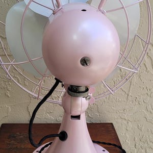 Puede incluir: Un ventilador el&eacute;ctrico de estilo vintage en un tono rosa suave. El ventilador presenta una jaula circular, tres aspas y una carcasa de motor redondeada. La base es rosa a juego, y el ventilador est&aacute; sobre una mesa de madera.