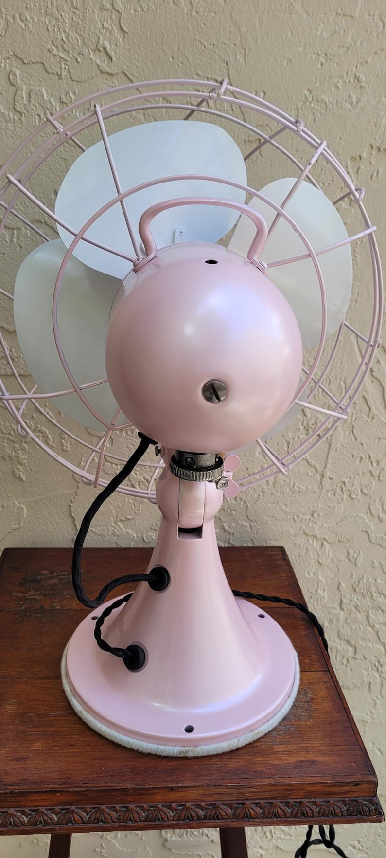 Puede incluir: Un ventilador el&eacute;ctrico de estilo vintage en un tono rosa suave. El ventilador presenta una jaula circular, tres aspas y una carcasa de motor redondeada. La base es rosa a juego, y el ventilador est&aacute; sobre una mesa de madera.