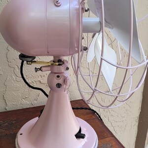Puede incluir: Un ventilador el&eacute;ctrico de estilo vintage en un tono rosa suave, con una carcasa de motor redondeada y una jaula protectora de alambre. Las aspas del ventilador son blancas. El ventilador est&aacute; montado sobre una base rosa a juego, sobre una mesa de madera.