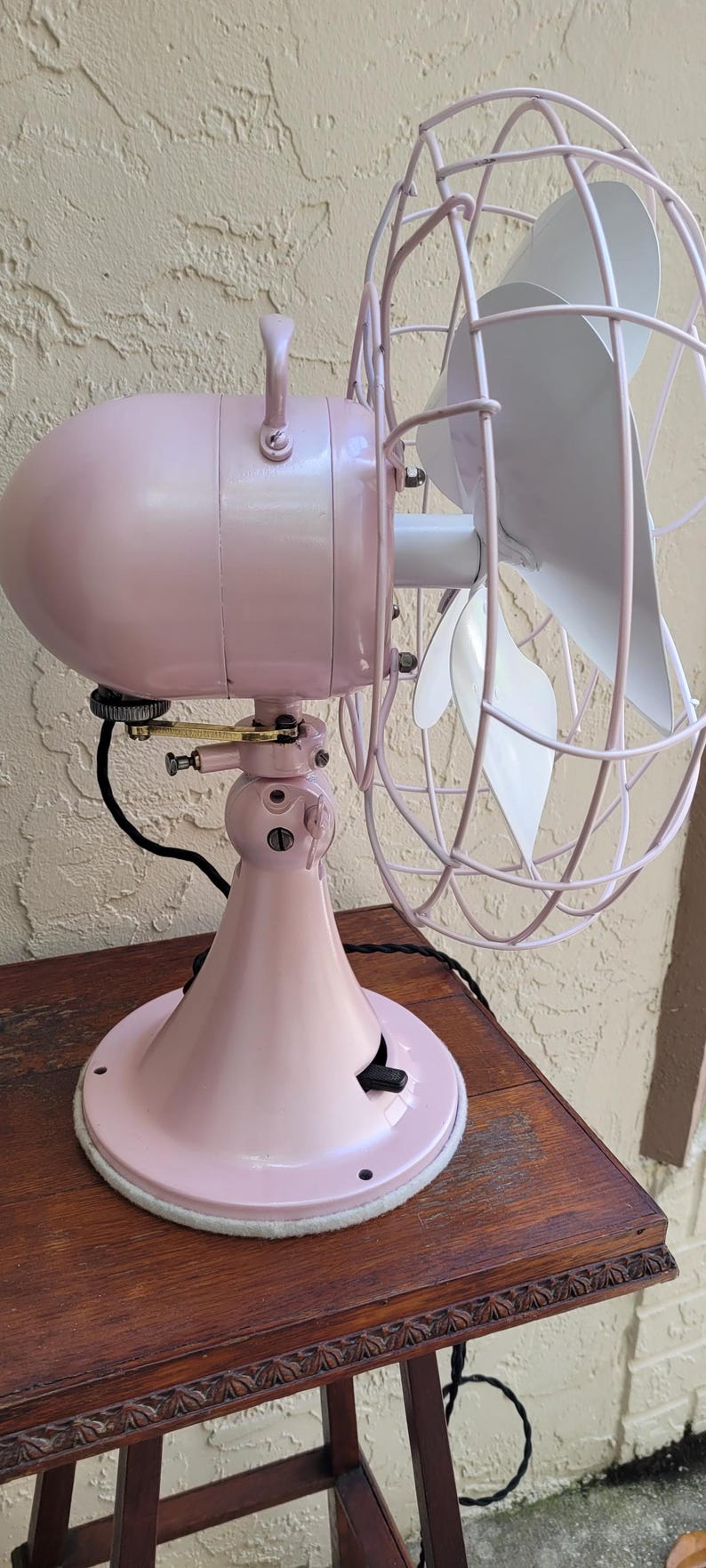 Puede incluir: Un ventilador el&eacute;ctrico de estilo vintage en un tono rosa suave, con una carcasa de motor redondeada y una jaula protectora de alambre. Las aspas del ventilador son blancas. El ventilador est&aacute; montado sobre una base rosa a juego, sobre una mesa de madera.
