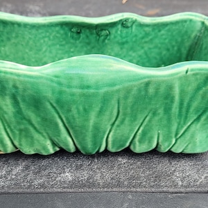 May include: A rectangular ceramic planter with a vibrant green glazed finish. The exterior features a textured, pleated design that mimics the appearance of gathered fabric, while the top rim has a wavy, undulating edge profile.