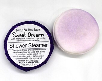 May include: A purple and white shower steamer from Bubble Bee Bath Treats. The label reads "Sweet Dreams" with mandarin, lavender, chamomile, neroli, and lily of the valley. The net weight is 1.5 oz.