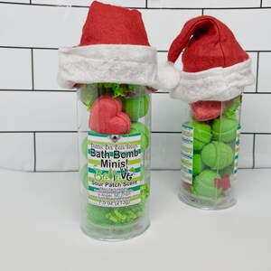 May include: Two clear plastic tubes filled with green bath bombs and red heart-shaped bath bombs, topped with a red Santa hat. The tubes have a label that reads "Bubble Bee Bath Treats Bath Bomb Minis! Believe Sour Patch Scent".