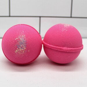 May include: Two bright pink bath bombs. One bath bomb has a band of glitter in shades of silver, blue, and gold. The other bath bomb has a band of glitter on top. The bath bombs are round and sit on a white surface.