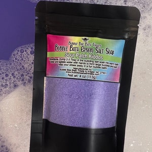 May include: A black pouch of Bubble Bath Epsom Salt Soak, labeled "Sour Patch Kiddos." The label is pink, green, and blue. The pouch contains a light purple powder. The net weight is 4 oz (113g). The product is surrounded by bubbles.