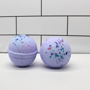 May include: Two lavender bath bombs with a speckled design of pink, blue, and teal. The bath bombs are spherical and have a seam around the middle. The background is a white tile wall.