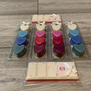 May include: A set of 18 heart-shaped wax melts in various shades of blue, pink, and red. The melts are arranged in three rows of six, with each row in a clear plastic container. There are also two wax melt bars in the image, one with a white top and a pink bottom, and the other with a white top and a white bottom. The wax melt bars have a label that says "Luxuries".