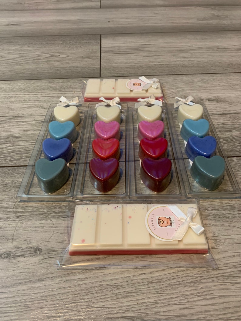 May include: A set of 18 heart-shaped wax melts in various shades of blue, pink, and red. The melts are arranged in three rows of six, with each row in a clear plastic container. There are also two wax melt bars in the image, one with a white top and a pink bottom, and the other with a white top and a white bottom. The wax melt bars have a label that says "Luxuries".