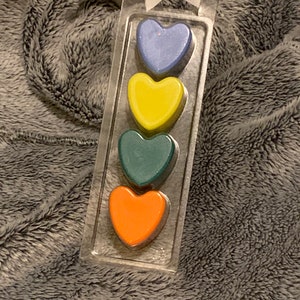 May include: A set of four heart-shaped wax melts in a clear plastic package. The melts are arranged vertically, with the top melt being blue, followed by yellow, green, and orange.