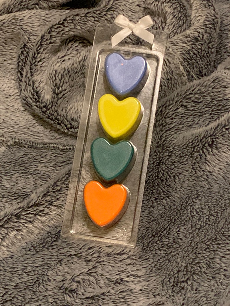 May include: A set of four heart-shaped wax melts in a clear plastic package. The melts are arranged vertically, with the top melt being blue, followed by yellow, green, and orange.