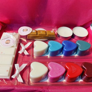May include: A pink gift box filled with scented wax melts. The box contains a variety of heart-shaped wax melts in blue, red, pink, and white. There are also two small white tea lights and a bar of wax melt in a brown wrapper.